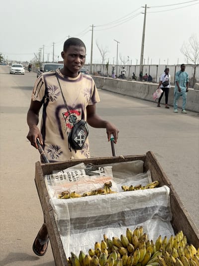The image captures an outdoor scene, likely a street or roadside market, under bright, possibly hazy, daylight. The main subject is a young man with dark skin and short, dark hair, positioned in the foreground, looking directly at the camera with a neutral expression. He is wearing a light-colored, short-sleeved t-shirt with a prominent dark-toned graphic or pattern, and a black shoulder bag with a white arrow logo (resembling the "OFF WHITE" brand) slung across his chest. He is holding two dark-colored handles or poles, indicating he is pushing or standing with a cart. His bare arms and dark-colored flip-flops are visible.
To the man's right (viewer's left), is a rustic wooden pushcart. It is heavily laden with numerous bunches of yellow, ripe bananas, which are visibly stacked both on top and within the cart. Some of the bananas inside the cart are partially covered by white, woven plastic sacks.
In the background, there's an asphalt road stretching into the distance. Several people are seen walking along the road, and others are gathered or seated on a lower level beyond a concrete barrier or wall that runs alongside the road. Tall utility poles with power lines punctuate the background against a light, hazy sky. Sparse, bare-branched trees are also visible in the distance. A white car is partially visible further down the road.
The visible text on one of the white plastic sacks in the cart includes: "A", "AZIO", "R", "CT", "ging", "GA S", "ALAGE", "IGERIA", "Line:", "748", "11", and "CS@".