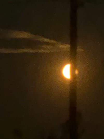 In the city of Baryshevo, Russia, a celestial event is captured. A half-illuminated moon, glowing with an intense orange hue, is visible against a dark, grainy sky. Wispy, pale clouds streak across the upper left portion of the frame. A dark, vertical structure, possibly a tower or antenna, bisects the image, partially obscuring the moon. A smaller, fainter celestial body or reflection is positioned to the right of the moon. The overall scene is dimly lit, with the primary light source being the moon itself.