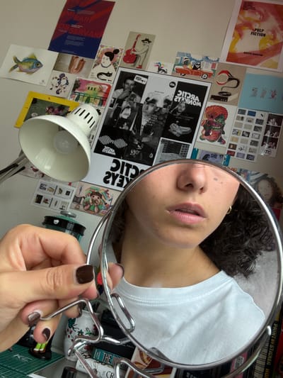 The scene, located in Jesús del Monte, Mexico, depicts an indoor environment focused on a self-reflection. In the foreground, a left hand with dark brown nail polish firmly grips a round mirror featuring an irregular metallic frame. The mirror reflects the lower portion of a person's face, showing slightly parted lips, the nose, and a small dark blemish on the right cheek. Dark, curly hair frames the face, and a gold hoop earring is visible on the right ear. The reflected torso shows a white t-shirt. To the immediate left of the mirror, a white articulated desk lamp with a bell-shaped shade is positioned, revealing a lit bulb.
The background consists of a light-colored wall heavily adorned with numerous posters, prints, and postcards. Visible decorations include a red and blue poster featuring the word "OBSERVAR," a watercolor illustration of a colorful fish, a print of a stylized woman holding a black cat, and a "Pulp Fiction" poster in red and yellow hues. Also present are an illustration of a skeleton driving a red car, an abstract design resembling a gramophone, and a large, black-and-white poster for "Arctic Monkeys" (text mirrored) displaying band members and a retro gaming device graphic. A grid of small photographic prints and a partial poster with the word "SHE" are also on the wall. Below the mirror, partially obscured, are a green cutting mat, a small figurine in green pants, a teal cylindrical container, and several books, including one titled "THE INK" and another "PAINT BY STICKER WORLD."