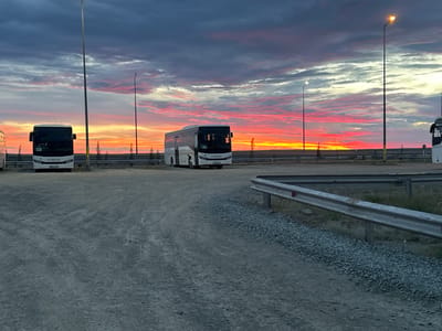 The image depicts a parking lot at sunset in Bozshakol, Kazakhstan. The sky is filled with dramatic clouds painted in vibrant hues of orange, pink, and purple, illuminated by the setting sun. Several white buses are parked in the lot, their headlights off. In the foreground, the ground is covered with gravel and dirt. A metal guardrail runs along the edge of the parking area. Streetlights are visible, some illuminated. The overall scene is one of quiet stillness under a colorful sky.