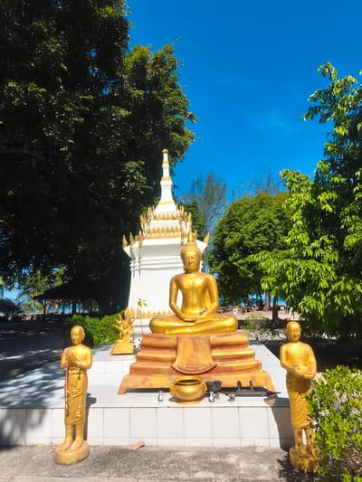 In Nathon, Thailand, a golden Buddha statue sits serenely on a raised, multi-tiered golden platform. The Buddha is depicted in a meditative pose, adorned with a golden crown. To the left and right of the main statue are two smaller golden statues of monks, also in prayerful poses. Behind the Buddha, a white stupa with golden decorative elements rises towards a clear blue sky. Lush green trees surround the statues and stupa, casting dappled shadows on the ground. In the background, hints of the sea and a pavilion suggest a coastal location. The scene is bathed in bright sunlight, creating a vibrant and peaceful atmosphere.