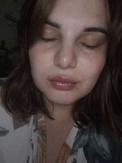 A close-up photograph depicts a woman with fair skin, dark brown hair, and closed eyes, lying on a patterned surface resembling bedding. Her lips are slightly parted, revealing a hint of pink. The background is dimly lit, providing minimal context. There is no information to suggest that the image was taken in the city of Амзя, Russia.