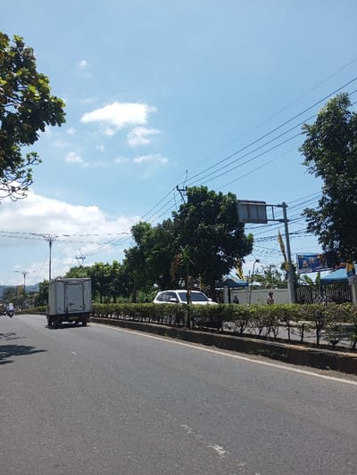 The image displays a daytime street scene under a clear blue sky with scattered clouds in Tagtse Tashidingkha, Bhutan. A two-lane asphalt road, divided by a raised median featuring trimmed green bushes, stretches into the background.
In the left lane, a white box truck, viewed from the rear, proceeds away from the camera. In the far distance on the left, other vehicles, including a motorcycle, are faintly visible. On the right side, behind the median and bushes, a white SUV is parked or stationary. Two human figures are observed on the right, near the SUV, positioned either standing or walking.
The roadside environment is characterized by numerous green trees, utility poles, and an array of overhead wires. On the right, a rectangular, unpopulated traffic sign structure stands. Further back on the right, a banner with indecipherable text and images, along with yellow flags, is visible near structures with blue and white awnings. Decorative streetlights are present on the left side of the road. Distant hills form the horizon in the background to the left.