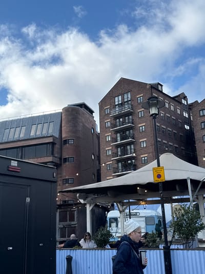 An upward shot captures a brick building with many windows, contrasted against a blue sky filled with fluffy clouds. A smaller, cylindrical tower-like brick structure adjoins the main building. A partially visible black trailer and an outdoor dining area with people present occupy the foreground. A yellow sign, possibly a parking restriction notice, is fixed to a pole.
