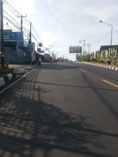 The image shows an asphalt road with a yellow line divider, likely in a city or town, due to the presence of buildings, streetlights, and utility poles. A person is walking on the left side of the road, near a blue building with a yellow fence. A billboard is visible further down the road. Motorbikes and other vehicles are present in the distance. The overall environment suggests an urban or semi-urban setting. However, the location of Phou Khoun, Laos cannot be verified based on the information in the image.