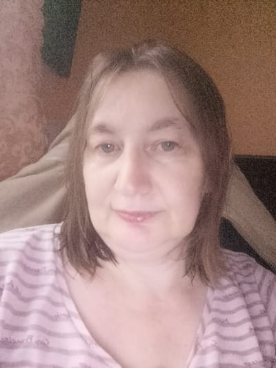 The image is a close-up, head-and-shoulders selfie of a middle-aged Caucasian woman with light brown hair and blue eyes. She is wearing a pink and white striped shirt. Her face is softly lit, and she has a slight smile. The background is out of focus and appears to be an indoor setting with a textured wall. There is no indication of the city of Berezynka, Ukraine, or any specific environmental details that would place the image in that location.