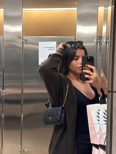 A female subject is captured in a mirror selfie inside what appears to be a public restroom in Lawrence Township, United States. She is seen from the waist up, holding a dark-colored smartphone with a black case and a yellow/orange logo (partially reading "MALLAH IS MY PLACE" in reverse) in her right hand, pointed towards the mirror. Her left hand is raised to her head, near dark sunglasses perched on her dark hair. She wears a dark, low-cut top under a dark brown, textured knit cardigan. A black quilted Chanel handbag with a gold chain strap is visible on her left side. To her right, several multi-colored shopping bags are reflected. The background consists of stainless steel stall doors, one of which displays a reversed white sign reading "TEMPORARILY CLOSED." Above the stalls, a light brown wall section with warm lighting is visible.