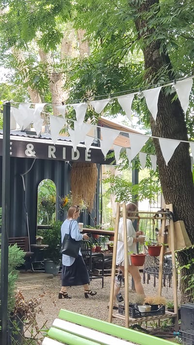 In Novaya Adygeya, Russia, an outdoor cafe setting is depicted, featuring a rustic and natural aesthetic. White triangular flags are strung overhead, and a large tree with lush green foliage dominates the right side of the frame. In the background, a building appears to be constructed from a shipping container, with the words "& RIDE" displayed prominently above the entrance. A woman wearing a light blue long-sleeved shirt and a dark skirt walks away from the building, carrying a black tote bag and a cup. She is wearing black heeled sandals. To her right, another person in a white top and light-colored shorts stands near a wooden display rack adorned with potted plants and decorative items. The ground is covered in gravel, and various plants, including small bushes and potted flowers, are visible throughout the scene, suggesting a garden-like atmosphere. In the foreground, a green bench is partially visible.