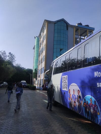The image depicts an outdoor scene under clear blue skies, located within the city of Hospital ward, Kenya. A multi-story building, a large passenger bus, and several individuals populate a paved area.
**Building:** A multi-story structure, estimated at six floors, dominates the background. Its exterior combines light-colored walls with large sections of blue-tinted reflective glass, particularly at its architectural corners. An adjacent section on the left features green-tinted glass. The roof line on the right displays a raised framework supporting two dark, cylindrical objects, likely water tanks.
**Bus:** A large, white passenger bus is parked on the paved surface, occupying the right midground. Its side is overlaid with a blue vinyl wrap featuring a prominent white QR code. Beneath the QR code and towards the rear, two circular graphics depict groups of young individuals, identified as athletes, with some holding sports equipment. Partial branding text, "Mo" and "SP," is visible on the wrap. The bus windows are tinted.
**Paved Area:** The ground consists of dark grey interlocking concrete blocks arranged in a repeating pattern.
**People:** Four individuals are clearly visible walking on the paved surface, generally moving from right to left across the frame.
1. A male in the foreground right, wearing a dark top, dark trousers, and a light-colored cap, with a beard, walks towards the left, carrying an object in his left hand.
2. A female in the mid-ground left, wearing a light-colored long-sleeved top and light trousers, holds papers in her left hand while walking towards the left.
3. A female further back on the left, wearing a dark top and light trousers, walks towards the left.
4. A fourth individual is partially visible in the background left, also walking.
No direct interactions are observed between these individuals.
**Environment:** Trees and green foliage line the far left of the scene and appear behind a cluster of parked vehicles in the distance. The scene is brightly lit by natural sunlight, casting visible shadows.