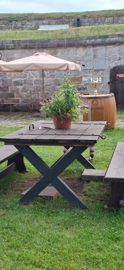 The image depicts an outdoor seating area, likely in a historical or fortress setting, with a focus on a wooden picnic table and benches. In the foreground, a dark wooden picnic table with an X-shaped base is set on a grassy area. A potted plant with green foliage and purple flowers sits on the table. To the right, a portion of a wooden bench is visible. In the background, a large beige umbrella is open, providing shade. Behind the umbrella, a stone wall forms the backdrop. A wooden barrel with metal bands and brass taps stands to the right of the umbrella, and to its right, a dark wooden door with a red sign is visible. The scene suggests a relaxed outdoor dining or resting spot within a fortified location, possibly a castle or old town. The presence of the stone wall and architecture hints at a location like the Kuressaare Castle in Kuressaare, Estonia, which is known for its well-preserved medieval fortress and surrounding historical context.