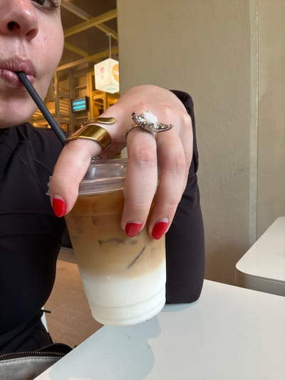 A person is holding an iced coffee in a plastic cup with a straw, with their lips visible on the straw. They are wearing two rings on their left hand: one silver with a white stone and one gold. The fingernails on their left hand are painted red. They are wearing a black shirt. The photo appears to have been taken indoors at a restaurant or café, with a hanging light fixture visible in the background. While the exact location within San Pedro Garza García, Mexico, is not discernible from the image, the presence of the hanging light and the style of the restaurant suggest it is a modern café or dining establishment within the city. A white table is partly visible on the right of the image, and a chair is also partly visible.