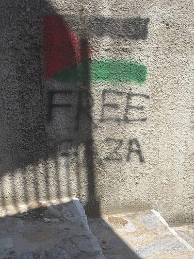 The image shows graffiti on a textured, light-colored wall. The graffiti includes three horizontal bands of color, red at the top, black in the middle, and green at the bottom, representing the Palestinian flag. Below the flag, the word "FREE" is stenciled in large black letters, followed by "ZA" in smaller black letters. A shadow cast by what appears to be a railing or fence falls diagonally across the wall and the graffiti, creating a striped pattern. The wall is situated outdoors, with a stone pathway or steps visible in the lower portion of the image. The setting appears to be an urban or semi-urban environment. The graffiti is in a public space, likely on an exterior wall.