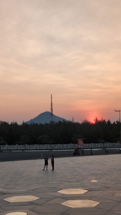 Here's a detailed description of the image:

**Location:** Unknown City, Vietnam

**Setting:** An outdoor area, possibly a plaza or public space, with a view of a mountain. The scene takes place during sunset.

**Foreground:**

*   A paved area featuring large, geometric tile patterns with inlayed shapes.
*   Two people are visible, walking on the paved area.
*   A line of parked motorcycles is near a walkway.

**Background:**

*   A large hill.
*   A tall tower is on top of the hill, likely a radio or communication tower.
*   A line of trees is in front of the hill.
*   The sky shows sunset. The sunset is orange and light pink.
*   A building or a wall is on the lower right.