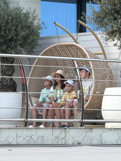 This media file depicts a group of people, likely children and an adult, resting in a hanging wicker chair on a balcony. The scene appears to be outdoors, with a portion of a building's exterior visible, featuring white siding and cracked marble-like accents. Green foliage from a nearby tree or plant is also present, partially obscuring the view.
The subjects are casually dressed in t-shirts and hats, suggesting a relaxed atmosphere. One individual, appearing to be an adult, is positioned at the back, leaning against the chair. In front, several younger individuals are seated, some looking at the camera, others looking away. They are wearing light-colored t-shirts and one is wearing a white baseball cap. The overall mood seems to be one of leisure and contentment.
There are no overt indications of a specific event, but the setting suggests a vacation or a warm-weather day. The lighting is bright, implying it is daytime, and the presence of light clothing suggests fair weather. The background includes a large blue pane, possibly a window or a painted surface, and the metallic structure of the balcony railing.
No discernible text is visible in the image, except for what might be a faint logo or print on one of the t-shirts, which is too indistinct to read. The location context provided indicates Yalta, Ukraine.