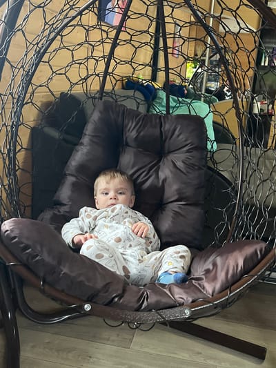 A baby is lying in a brown hanging chair indoors, likely in a home. The chair has a woven metal frame and a large, padded cushion. The baby is wearing a white patterned pajama suit and blue socks. The background shows a wooden floor and glimpses of items stored on shelves. The image was taken in the city of Protasy, Russia.