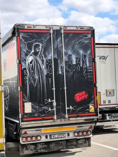 The image shows the rear doors of a white semi-trailer truck that has been painted with a mural. The mural depicts a stylized image of a man holding a gun, standing in front of a cityscape with lightning in the background. The words "SIN CITY" are painted in red lettering on the mural. The truck is parked on asphalt, likely in a lot or on a road. Another semi-trailer truck, white with a red stripe, is parked to the right, with partial markings visible including "RDV Transports" and speed limit signs for 60 and 80. The license plate of the first truck is visible and reads "KWI 695PK". The location in the city of Briis-sous-Forges, France is not specified in the image.