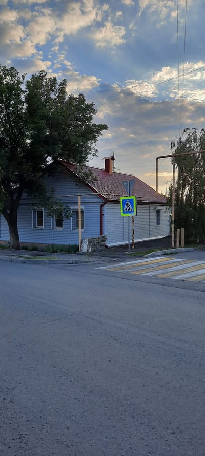 The image depicts a street scene in Shakhty City District, Russia, on a cloudy day with sunlight filtering through. In the foreground, a paved road with visible cracks occupies the lower half of the frame. A pedestrian crossing with yellow and white stripes is prominent on the right side of the road. Adjacent to the crossing, a blue and yellow pedestrian crossing sign is mounted on a post.
Behind the road and crossing is a light blue, single-story house with a red tiled roof. The house has several windows and a visible chimney. A large, mature tree with dark green foliage stands to the left of the house. The background features more trees and utility poles with wires. The sky is filled with a mix of white and gray clouds, with shafts of sunlight breaking through, creating a dramatic effect. The overall impression is a quiet, residential street bathed in the late afternoon or early morning light.