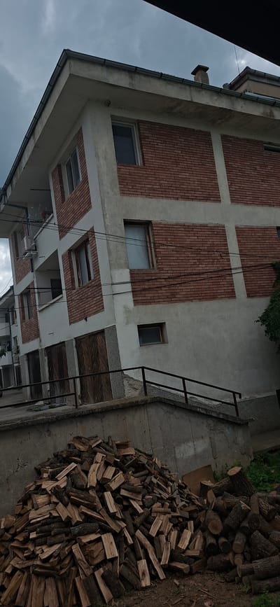 The image shows an outdoor scene, rotated 90 degrees clockwise from its natural upright orientation.

The main subject is a multi-story building facade, which occupies the left and central-left portion of the frame. The building features a light grey, possibly concrete or stucco, exterior interspersed with large, rectangular panels of red brickwork arranged in a grid pattern. Several windows are visible; some reflect the grey sky, while others appear as dark openings. Dark rain gutters are present along the visible edges of the building's rooflines. Black cables or wires hang vertically down the building's side. Towards the bottom, an overhanging section of the building contains further windows or skylights, also framed by brickwork.

To the right of the building, a substantial pile of chopped firewood, consisting of logs and smaller pieces, is stacked on an uneven surface of concrete and dirt. A black metal, ladder-like structure stands on the concrete ground, extending upwards towards the building. Some green foliage is visible in the upper right corner of the frame.

The sky, visible towards the top-left, is uniformly grey and cloudy, indicating overcast weather conditions. The setting appears to be an exterior utility area or backyard of a building, located in Strazhitsa, Bulgaria. No people or visible text are present in the image. The scene is static, capturing a functional outdoor environment.