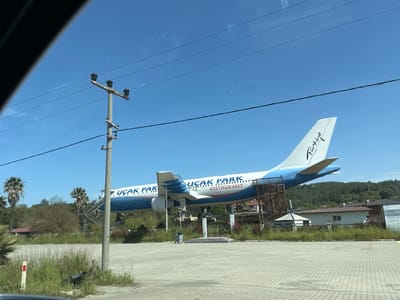 The image shows a decommissioned aircraft repurposed as a restaurant, "Uçak Park Restaurant," located in Ula, Turkey. The plane is painted in white and blue, with the word "Türkiye" written on the tail fin. The plane is elevated off the ground and has a stairway to enter. A utility pole stands in the foreground, and there is vegetation and a building in the background. A parking lot is visible in the foreground.