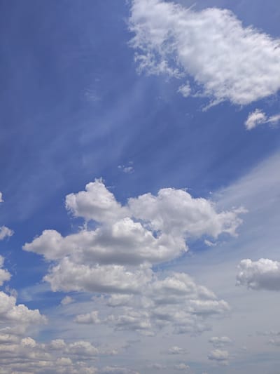 The image shows a wide-angle view of a bright blue sky filled with a variety of white and gray clouds. The clouds range from wispy, thin cirrus clouds stretched across the upper portion of the frame, to larger, more defined cumulus clouds scattered throughout the midground and foreground. The lighting suggests it is daytime, with the sun likely positioned out of frame, illuminating the clouds from above. There are no people, buildings, or any other discernable elements of a city or landscape visible in the image. The focus is entirely on the atmospheric conditions.