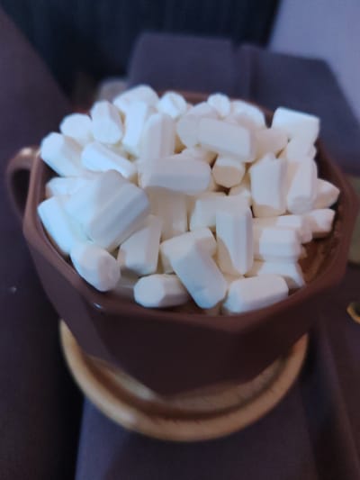 A close-up, slightly out-of-focus shot shows a brown faceted mug filled with white mini marshmallows. The mug is sitting on a light brown saucer. The background is dark and blurry, suggesting an indoor setting with low lighting. There are no people visible in the image. The scene is static and no actions are taking place. There is no indication of the location in Polatsk, Belarus.