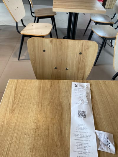 This is a high-angle, medium shot of a wooden table with a receipt on it. The table is light brown with a wood grain texture. In the background, there are several wooden chairs with black metal legs. The chairs are arranged around a similar wooden table. The overall scene suggests a casual dining or cafe environment. The receipt is white and partially unrolled, showing printed text and a QR code. The text on the receipt indicates a transaction at KFC, with details about items purchased and promotional offers. The background is slightly out of focus, emphasizing the table and receipt in the foreground. No people are visible in the image.