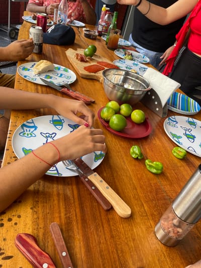 This detailed image depicts an outdoor dining scene in Coatzacoalcos, Mexico, featuring a gathering of multiple individuals around a polished wooden table. At least four people are partially visible, identified by various hands and arms. In the upper left quadrant, a person's arm, wearing a patterned shirt, holds a silver beer can, next to which rests a dark blue baseball cap. On the right side, an individual is partially visible, wearing a black top with a red garment draped over the shoulder. In the foreground, two hands are positioning a fork and knife with wooden handles onto a white oval plate decorated with blue and green fish patterns. A red string bracelet is visible on the left wrist.
The table is extensively covered with food, beverages, and dining accessories. A wooden cutting board prominently features slices of cured ham and round slices of pepperoni or chorizo. Adjacent to this, a red oval tray contains five green limes. A stainless steel mixing bowl sits near the limes, and three individual, irregular green peppers, consistent with habaneros, are placed directly on the wooden surface. Other food items include a split bread roll on another fish-patterned plate and a small dish containing an avocado-based dip, likely guacamole.
Beverages and condiments include additional canned beers, a clear glass bottle, a red squeeze bottle (ketchup), and a yellow squeeze bottle (hot sauce). A tall glass filled with a dark brown liquid is also present. The table is set with multiple white oval plates featuring fish designs and a stack of striped blue and white oval plates. Cutlery, including several knives and forks with distinctive wooden handles, is distributed. A stainless steel napkin holder with white paper napkins and a stainless steel salt grinder containing pink Himalayan salt are also visible on the table. The background shows a partial view of an outdoor setting, including what appears to be a brick wall.