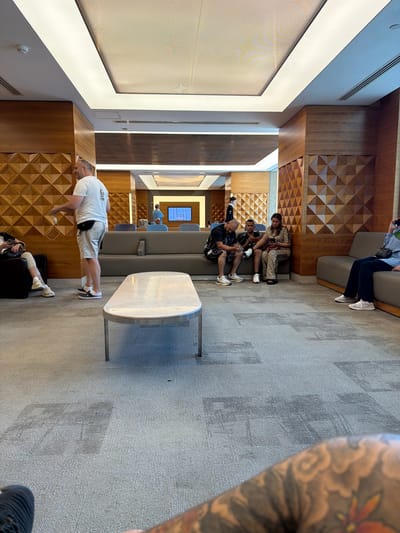 A man in a white t-shirt and shorts is doing a plank exercise in a lounge area. He is positioned on a grey carpeted floor with a light-colored, oval-shaped table in front of him. Behind him, a wall features a geometric wooden paneling design. Further back, several people are seated on a grey couch. One person is on their phone, and two others are talking. Another person in the background is walking. The room appears to be an indoor waiting area, possibly an airport lounge, given the presence of seating, a television screen displaying a blue image, and the overall sophisticated design. The lighting suggests it is daytime.
