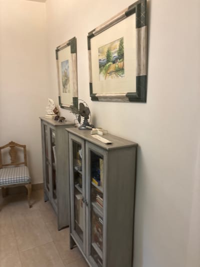 The scene is an interior. Two framed paintings hang on a light-colored wall above a gray, glass-fronted cabinet. On top of the cabinet sits a small vintage fan, books, and decorative items. To the side is a chair with a plaid cushion. The location is not apparent from the image. No people, interactions, or actions are visible.
