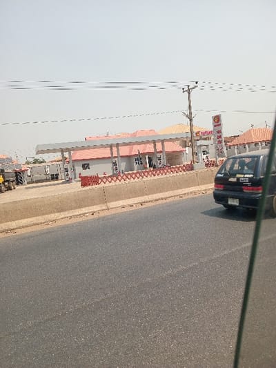 An image captures a daytime scene along a paved asphalt road in AlakIa, Nigeria, offering a view of a "TOTAL" branded fuel service station. In the immediate foreground, a dark-colored sedan with a visible license plate is observed driving away from the viewer on the road. A concrete barrier separates the road from the service station's entrance. The service station features a prominent white canopy covering multiple fuel dispensers. Behind the canopy, a light-colored building with a distinctive red tiled roof is visible, likely housing the station's amenities. A tall, red and white "TOTAL" sign stands to the right, displaying the brand name and additional text that appears to include "PAYMENT." A red decorative fence delineates the front perimeter of the station. To the left within the station compound, yellow construction equipment and several white container-like structures or trucks are parked. Further in the background, additional buildings with red and beige tiled roofs are partially visible under a bright, hazy sky. Overhead power lines span the upper portion of the frame, supported by a wooden utility pole. No individuals are clearly discernible.