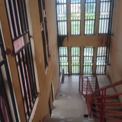 The image depicts a dimly lit interior staircase with barred windows. The walls appear to be a pale, yellowish-brown color and show signs of wear and tear. Several large windows, each with thick black bars, line the upper portion of the staircase. Through the windows, glimpses of greenery and other buildings can be seen, suggesting an outdoor environment beyond the structure. The staircase itself has a red metal railing and appears to be made of concrete or tile. A few objects are scattered on the floor, including what looks like a fallen bottle or container. The scene is observed from an upper level, looking down the staircase. The image is too blurry to identify any people or specific actions taking place. The overall impression is of an aged and somewhat neglected building.