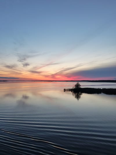 The image depicts a serene sunset over a body of water in Kosino, Russia. The sky is painted with hues of pink, orange, and blue, with wisps of clouds stretching across the horizon. The calm water reflects the vibrant colors of the sky, creating a mirror-like effect. In the middle ground, a small island with a lone tree and reeds stands in the water. The foreground shows gentle ripples on the surface of the water, suggesting a slight breeze. The overall impression is one of tranquility and natural beauty at the close of day.