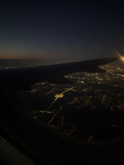 The image displays an aerial night view from an aircraft, presenting a dark sky transitioning to a faint horizon gradient in the upper left. A significant portion of a dark airplane wing extends diagonally from the mid-left towards the upper right, silhouetted against the dark sky and illuminated ground. A concentrated, bright light source, likely from the aircraft, emanates from the far right tip of the wing. The lower two-thirds of the frame reveal a vast urban landscape characterized by numerous artificial lights forming intricate patterns below. These lights outline roads, infrastructure, and developed areas. A prominent, brightly illuminated, irregularly shaped complex is visible towards the center-left of the lit ground, connected by a distinct, brightly lit linear feature, consistent with a major road or highway. The curved interior edge of an aircraft window frame is visible along the bottom and left sides of the image. No individuals, explicit actions, or interactions are discernible. The depicted location is the city of Korzeniówka, Poland, observed from an altitude during darkness.