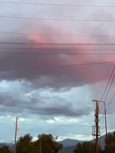 The image depicts an outdoor scene with a dramatic sky. The sky is filled with thick clouds, some of which are illuminated with a soft pink hue, suggesting either sunset or sunrise. In the foreground, several power lines stretch horizontally across the frame. To the right, a wooden utility pole stands, supporting electrical equipment and a street light. Another wooden pole is visible to the left, also with insulators for wires. Below the sky and power lines, the tops of trees and distant mountains can be seen, indicating a rural or semi-rural setting. The overall atmosphere is somber yet visually striking due to the interplay of light and clouds. The location is identified as Sarymoldayev Rural District, Kazakhstan.