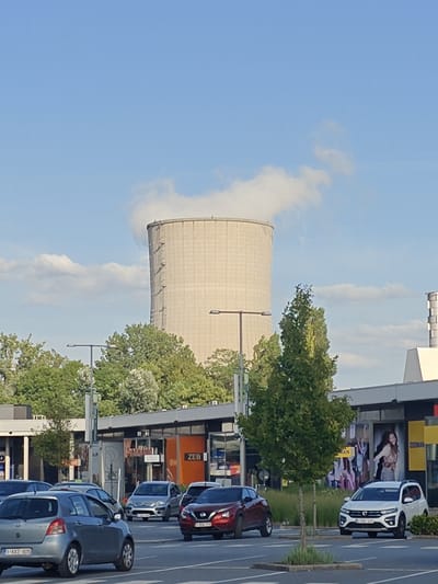The image depicts a scene in Sint-Pieters-Leeuw, Belgium, under a clear blue sky with scattered clouds, suggesting it's daytime. The focal point of the image is a large, cylindrical cooling tower, emitting a plume of white steam. This suggests an industrial setting, likely a power plant. In the foreground, several cars are parked in a lot, and some are on a road, indicating a public or commercial area.
The lower half of the image shows a row of buildings with storefronts. One of these stores has a prominent orange sign with the white text "ZEB" visible. The presence of shops and a parking lot suggests a commercial zone, possibly a shopping center or retail park.
The cars in the foreground include a silver hatchback, a red SUV, and a white SUV. The license plate on the silver car reads "1 KXZ-905". The white SUV has a license plate that is partially visible, appearing to start with "1-AZC-".
The overall atmosphere is bright and casual, typical of a day with good weather. The cooling tower dominates the background, providing a distinct landmark, while the foreground elements of cars and shops offer everyday context. The scene appears to be a snapshot of a functional, populated area with both industrial and commercial aspects.