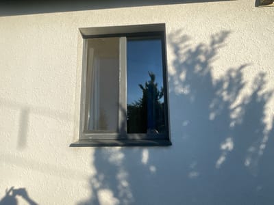 This is a medium-shot, eye-level photograph of a window on the exterior of a textured, off-white building. The window is rectangular with a dark grey frame. It is a sliding window with two panes, one of which appears to be a screen. The right pane is reflective, showing a dark blue sky and the silhouettes of trees. The wall itself has a rough, stucco-like texture and is partially covered by dappled shadows that suggest sunlight filtering through tree leaves. The shadows are cast on the right side of the wall and extend across the window. A small, rectangular vent is visible in the upper right corner of the building's exterior wall. The lighting suggests it is daytime, with the sun casting strong shadows. The location is identified as a rural area in Belarus. No people or explicit activities are depicted, but the presence of the screen suggests it might be a warmer season. The overall scene is calm and ordinary, focusing on architectural details and the interplay of light and shadow.
