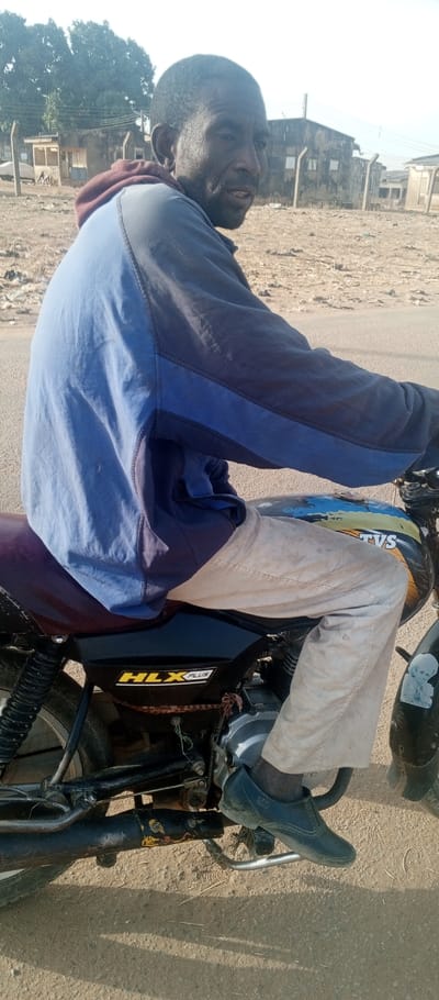 A male individual with dark skin, short dark hair, and a short beard is depicted seated on a black motorcycle in an outdoor setting in Tunga Mekidi, Nigeria. He is wearing a two-toned blue hooded jacket with a visible reddish-brown hood, light-colored trousers, and dark, low-cut shoes. His right foot rests on a footpeg. The motorcycle features a dark reddish-brown seat, a discolored fuel tank with blue, yellow, and possibly white patches bearing "TVS" branding, and a black side panel marked with "HLX PLUS" in yellow lettering. A sticker depicting a cartoon child's face is affixed to the front mudguard. The exhaust pipe is black with yellow and orange discoloration. The man is looking directly towards the viewer with his mouth slightly open. The ground beneath the motorcycle is an unpaved, light brown surface. In the background, several weathered buildings with flat or low-pitched roofs are visible, some partially obscured by fencing. Green foliage from trees is present on the far left. The sky is bright and appears hazy or overcast.