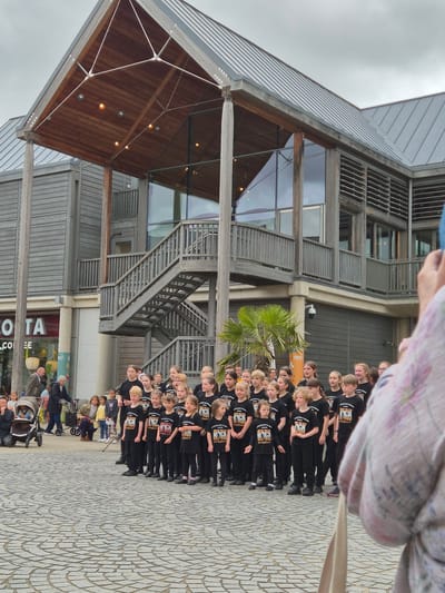 A group of approximately 25 children and teenagers, mostly girls, are arranged in several rows, standing formally on a patterned stone plaza. They are uniformly dressed in black t-shirts with white lettering that reads "ROCK YOU" and "young@sport," facing towards the right side of the frame. In the extreme right foreground, the out-of-focus shoulder and arm of an individual holding a phone or camera are visible, indicating they are photographing the group.

The backdrop is a modern, multi-story building identified as the Arc in Bury St Edmunds, West Suffolk, United Kingdom. The building features an exterior clad with light grey wooden slats, large glass windows, and a prominent, expansive wooden canopy over an entrance, supported by a triangular steel framework and featuring recessed lighting. A substantial external wooden staircase with railings ascends to an upper level on the left side of the building. To the far left, a "Costa" coffee shop occupies the ground floor, with pedestrians visible nearby. The plaza surface is paved with light grey, irregularly shaped stones laid in radiating and concentric patterns. A small palm tree is situated to the left of the children. The sky is overcast.