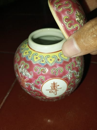 A close-up, high-angle image depicts a small, decorative ceramic jar with its lid partially open, located in the city of Longmiang, Laos. The jar possesses a spherical body, predominantly dark pink/red, with a white rim encircling its opening. A yellow band, featuring blue-outlined heart-shaped or scroll motifs, is positioned below the rim. The main body of the jar is decorated with swirling green and yellow patterns, along with white cartouches. A prominent white circular medallion on the front displays a red Chinese character, '寿' (shòu), signifying longevity. To the left, an elongated white ornate scrollwork design is visible. A human hand, with light brown skin and visible thumb and index finger, grips and elevates the matching ceramic lid. The lid shares the dark pink/red background with yellow/green scrollwork and a central white floral pattern. The jar rests on a dark reddish-brown surface, while the background is dark and out of focus.