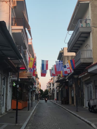 The image shows a narrow cobblestone street lined with buildings on both sides, decorated with numerous national flags strung across the street. The buildings appear to be multi-story structures with balconies and shops on the ground floor. On the left side of the street, there is an awning covering a shopfront, and an orange display unit. On the right side, there are shops with glass windows. A moped with a rider is visible in the distance, moving down the street. The sky is clear and appears to be either early morning or late evening, with a soft, warm light illuminating the scene. The location is identified as Alexandroupolis, Greece, based on the text on a sign visible on the left side of the street, which reads "ΦΡΟΥΤΑ - ΛΑΧΑΝΙΚΑ ΠΑΡΑΣΚΕΥΟΠΟΥΛΟΥ" (Fruits - Vegetables Paraskevopoulou).