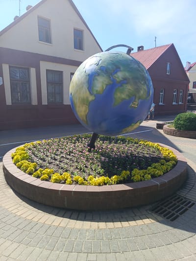 The image captures a street view of a large, terrestrial globe monument situated in a circular flower bed. The globe features painted continents and oceans, with city names in Cyrillic script visible, including Moscow and Beijing. The flower bed is bordered by a brick wall and is filled with soil, with yellow flowers lining the outer edge and smaller pink flowers in the center. The monument stands on a paved area with interlocking bricks forming a radial pattern. In the background, there are two buildings with tiled roofs and light-colored facades. The left building is cream-colored with dark brown trim and visible windows, while the right building is dark red with white-trimmed windows. A person wearing a light blue shirt and shorts is visible standing near the right building. The scene is illuminated by sunlight, casting shadows on the ground. This monument is located in the city of Kobryn, Belarus, likely in a public square or pedestrian area.