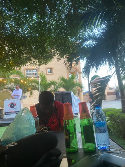 **Scene:** Outdoor gathering in Alagba, Nigeria.
**People:** A boy wearing a red shirt is visible in the foreground. A man in white is standing near a DJ setup.
**Objects:** A table holds a bottle of water, two green bottles with plastic cups, and a plastic bag. There is a DJ setup with speakers. The backdrop features palm trees, a building with windows, and string lights.
**Environment:** Trees and foliage surround the scene. The time of day appears to be late afternoon or early evening.