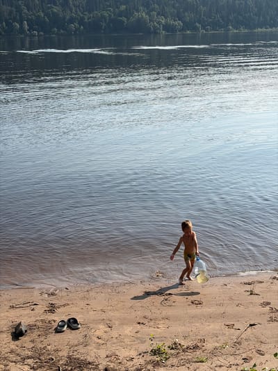 A young boy, wearing yellow swim trunks, stands on a sandy shore near a body of water. He is holding a large, partially filled plastic bottle in his right hand. The boy is looking down and appears to be in motion, with his left leg slightly extended. The water is calm with gentle ripples, and a line of trees forms a dense forest on the distant shore. The sand is light brown and shows evidence of scattered debris, including twigs and some dried vegetation. A pair of dark sandals are visible on the sand to the left of the boy. The scene appears to be a natural outdoor environment, possibly a riverbank or lake shore, in Ol'khovskoye, Russia. The lighting suggests it is daytime, with the sun casting shadows on the sand.