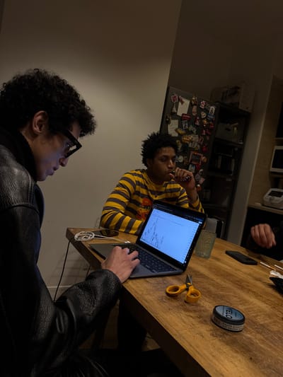The scene depicts an indoor gathering in Saint-Denis, France, centered around a wooden table. Two males are prominently featured, with a third person partially visible.

On the left, a male with dark, curly hair and dark-rimmed glasses is seated, wearing a black jacket (likely leather). He is actively engaged with a silver-colored laptop, his right hand positioned over the keyboard/trackpad. The laptop screen displays a financial candlestick chart. Next to the laptop, a white charging cable is coiled, and a black smartphone lies face down.

To the right, a second male with dark, curly hair and a yellow and dark striped shirt is seated, holding a black and red vaping device to his mouth. He is looking towards the laptop screen. A textured clear glass, partially filled with liquid, sits near him.

On the table in the foreground, yellow-handled scissors and a blue and white cylindrical container labeled "VELO" are present. Towards the far right, a hand is visible, and several cigarettes are on the table or in an adjacent receptacle.

The background shows a domestic environment, including a refrigerator adorned with numerous magnets and notes, and a shelving unit with various kitchen appliances, indicating a kitchen or dining area. The lighting is warm and somewhat dim.