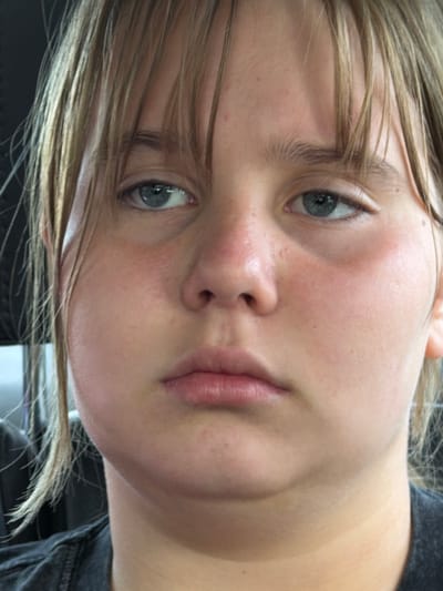 Close-up portrait of a fair-skinned person with light brown hair and blue eyes. The person is facing slightly to the left of the frame and looking at the camera. The background is out of focus, revealing only a gray or black fabric, possibly clothing. No discernible details indicate a location in Mshaga, Russia.