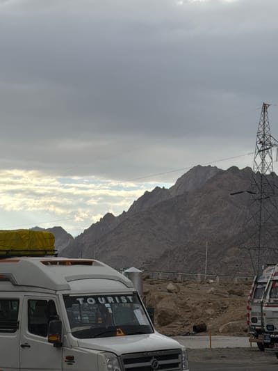 The image shows a cloudy sky over a mountainous landscape. In the foreground, a white tourist van is parked. The van has a yellow tarp on its roof and the word "TOURIST" is visible on its windshield. To the right of the van, part of a white truck with red and blue stripes is visible. The background consists of rocky mountains and a power pylon. The location is identified as Chumkar, India.