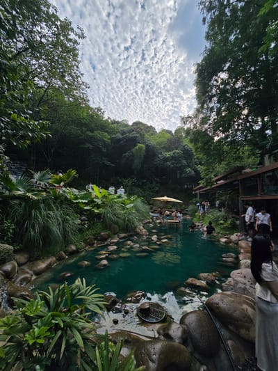 The image depicts an outdoor natural hot spring in Xiaoshan District, China, surrounded by lush greenery and rocky terrain. The spring water is a vibrant turquoise color. Several people are seen enjoying the hot spring, with some wading in the water and others relaxing on the banks. A yellow umbrella provides shade near a wooden structure in the water. The background is filled with dense trees and a sky with scattered clouds. The scene conveys a sense of tranquility and natural beauty.