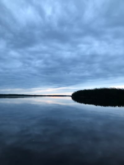 The image depicts a tranquil, still lake with a dark, treed shoreline visible in the distance. The sky is overcast with heavy, blue-gray clouds. The water perfectly mirrors the sky and the distant landscape, creating a symmetrical and serene scene. The lighting suggests either early morning or late evening. The location is identified as Svirstroy, Russia. There are no people or explicit actions captured in the image.