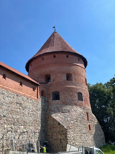 The image shows a close-up, low-angle view of the Trakai Island Castle in Trakai, Lithuania. The focus is on a circular brick tower with a conical red-tiled roof, topped with a metal spire flying a dark flag. The tower is constructed from red brick and features several arched windows, some of which appear dark. The lower portion of the tower and the adjacent wall are made of rough-hewn stones. To the left of the tower, a section of a brick wall with a red-tiled roof runs horizontally, also constructed with stone at its base and featuring small rectangular windows. The wall is undergoing some form of construction or renovation, with scaffolding and construction materials visible at its base. Two individuals, dressed in high-visibility safety vests and hard hats, are working near the scaffolding. One person, wearing a yellow vest and blue helmet, is standing and appears to be working on the scaffolding. Another person, wearing a bright yellow vest, is bent over near the ground. In the background, to the right of the tower, is a dense canopy of green trees. The sky is clear and bright blue.