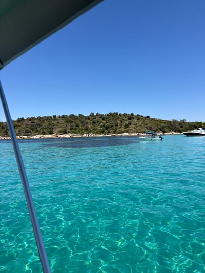A bright, clear day is depicted with a vast expanse of turquoise water in the foreground. The water exhibits a shimmering effect due to sunlight filtering through it, revealing the sandy bottom in some areas. A gray pole, likely part of a boat's structure, runs vertically along the left side of the frame. In the mid-ground, a small white motorboat with a blue canopy is visible, carrying at least two individuals. Further to the right, a larger white motorboat with a dark hull is anchored. The background features a verdant, hilly island densely covered with trees and shrubs. The shoreline is rocky, with patches of lighter colored sand. A clear blue sky stretches across the top of the image. The scene evokes a sense of tranquility and recreation, suggesting a location in a coastal area, possibly Greece, known for its clear waters and islands.